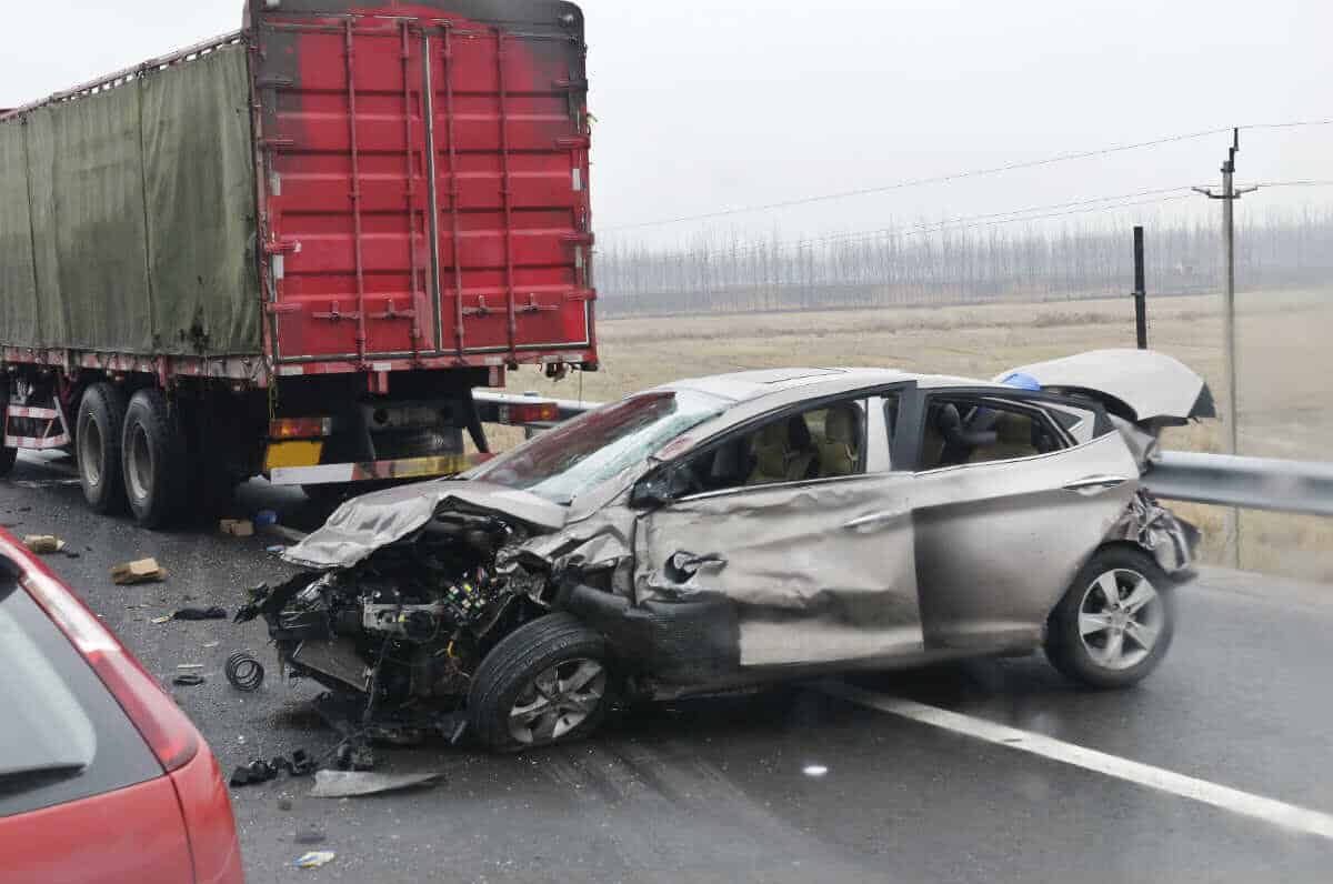 A severely damaged silver car is stopped in front of a large red truck after an auto accident on a wet, rural Kentucky road. The car's front end is crushed as insurance adjusters arrive to assess the scene.