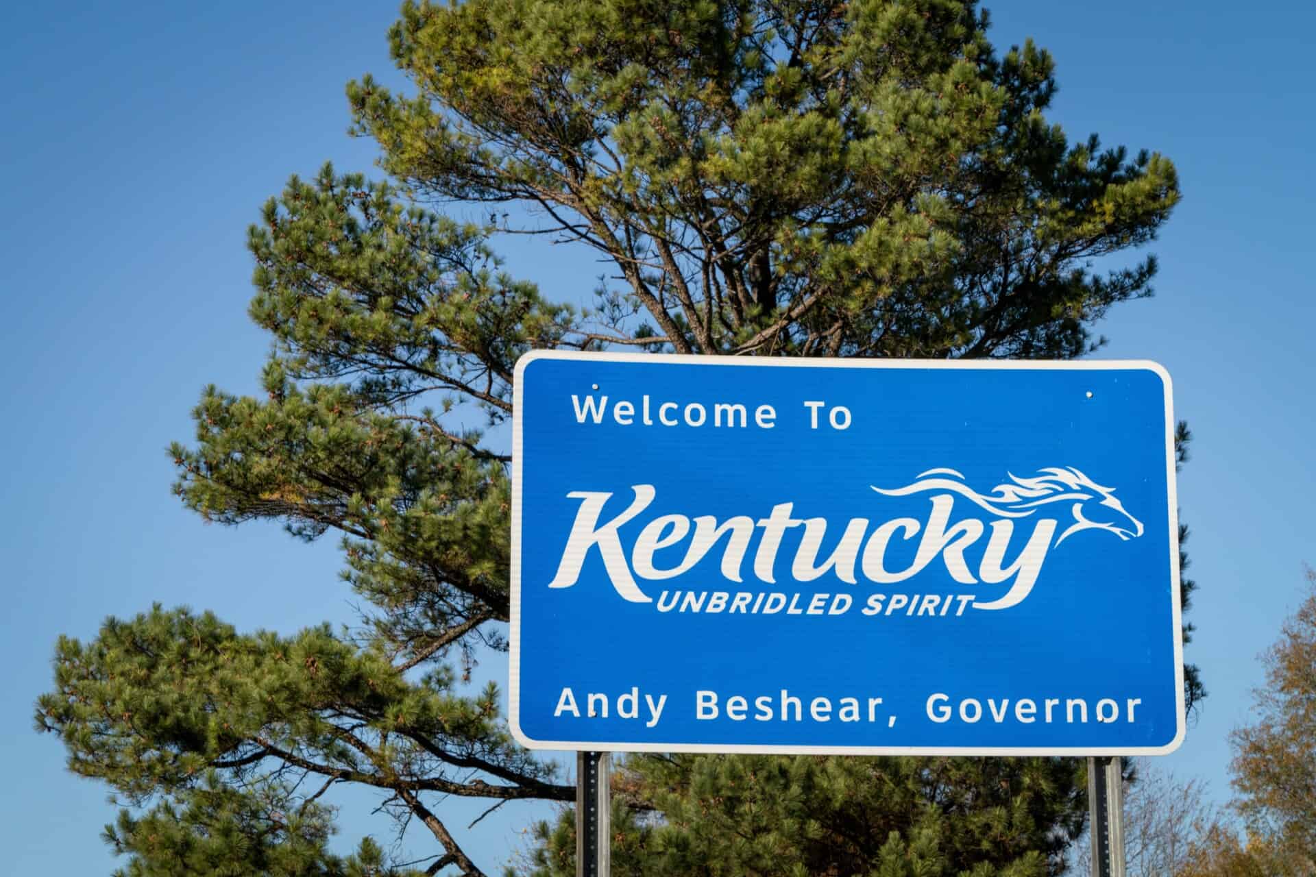 Blue roadside sign reads "Welcome to Kentucky Unbridled Spirit, Andy Beshear, Governor" with trees and blue sky in the background—reminding travelers of local laws like the Kentucky two-year rule legal deadline.
