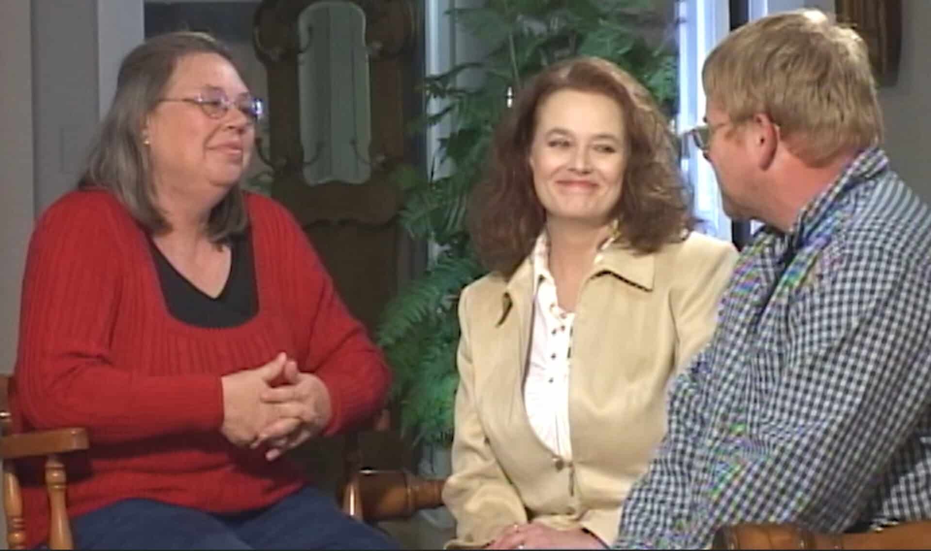 Three adults sit indoors, conversing about the Kentucky two-year rule. One woman in a red sweater faces a smiling woman in a tan jacket and a man in glasses and a checkered shirt.