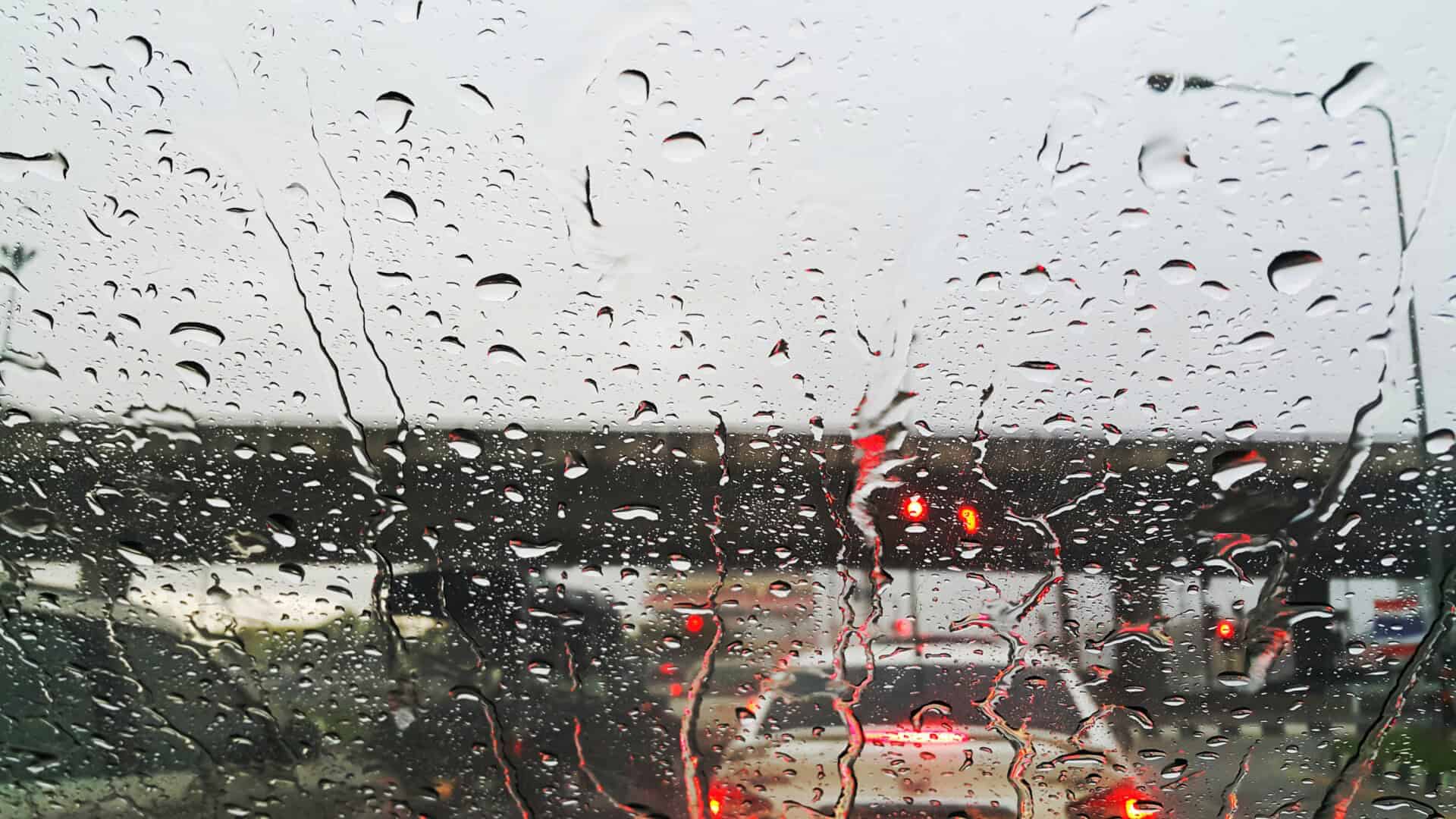 Rain droplets cover a car windshield, blurring the view of cars and traffic lights at a stop, as changing weather conditions create an overcast sky in the background.