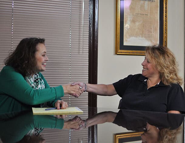 Two women sit across a desk from each other, smiling and shaking hands in an office setting with a framed document on the wall, perhaps discussing medical bills after a recent fender bender.