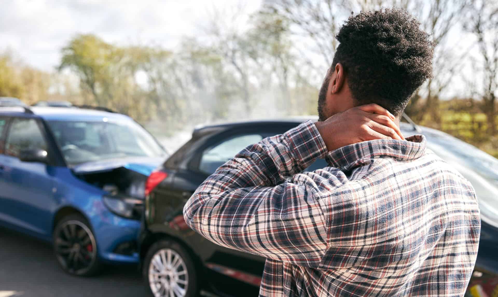 A person holding their neck stands near two cars involved in a fender bender on a road during the day, possibly facing medical bills after the minor Kentucky crash.