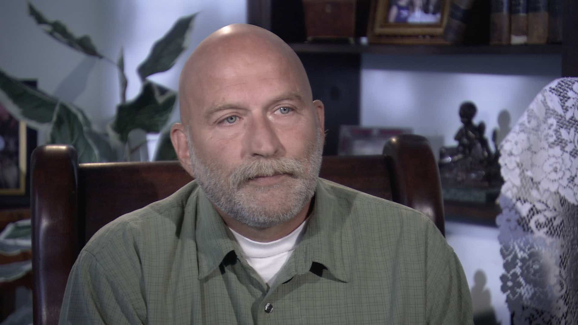 A bald man with a beard wearing a green shirt sits indoors at home, with shelves, books, and plants in the background.