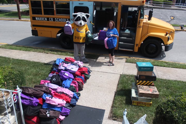 A person and the Warren County raccoon mascot stand by a school bus next to piles of colorful backpacks and stacked boxes on a sidewalk.