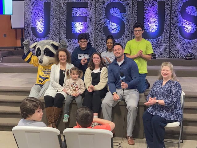 A group of people, including a raccoon mascot, sit and stand on steps in front of a large "JESUS" sign in a church setting. Some are smiling, and one person holds a microphone at the Barren County event.