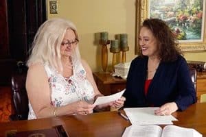 Two women sitting at a table review legal documents together, smiling. Open books and papers are spread out in front of them in a home office setting.