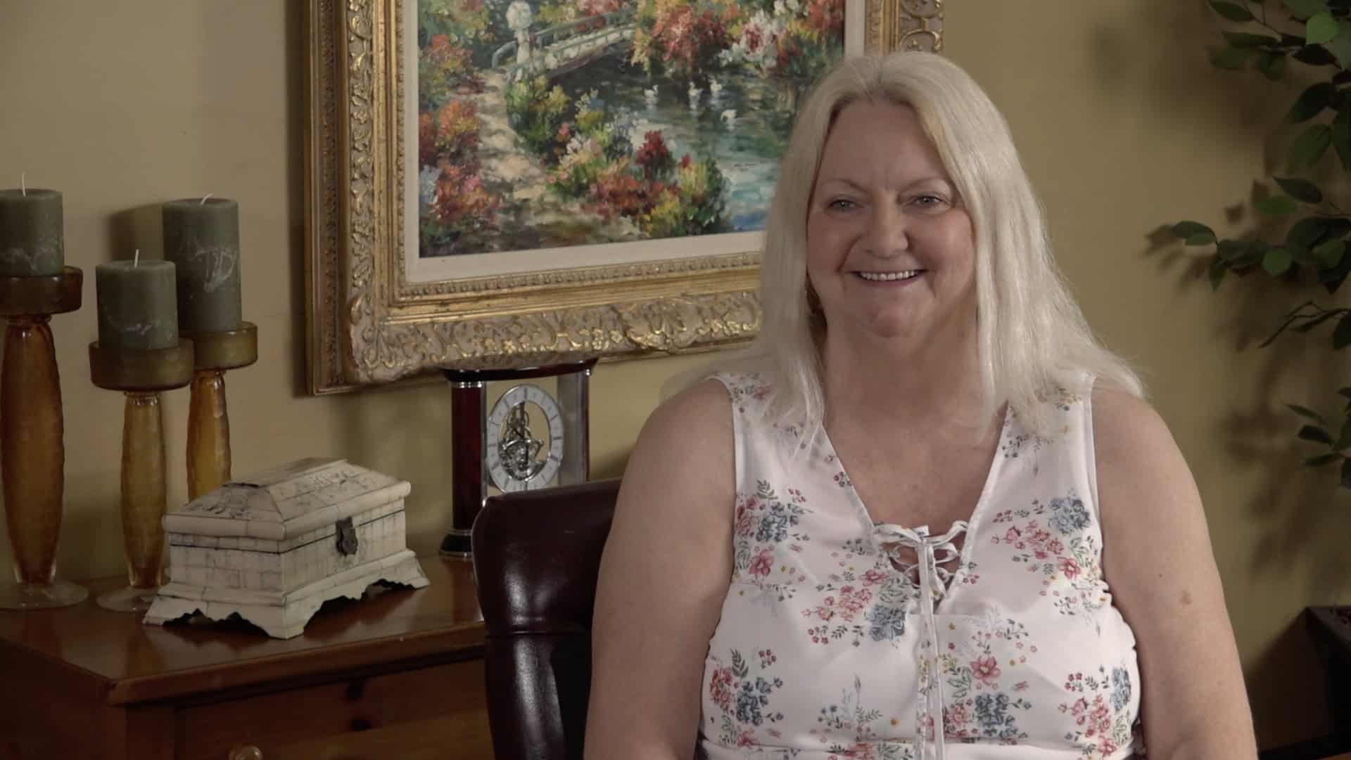 A woman with long blonde hair, wearing a sleeveless floral blouse, sits smiling at home in a room with a painting, candles, and a clock on a wooden table behind her.