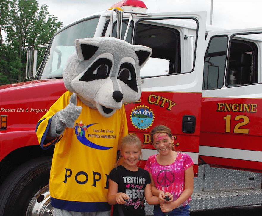 A person in a raccoon mascot costume poses with two smiling girls in front of a red Barren County fire truck labeled "Engine 12" on a cloudy day.