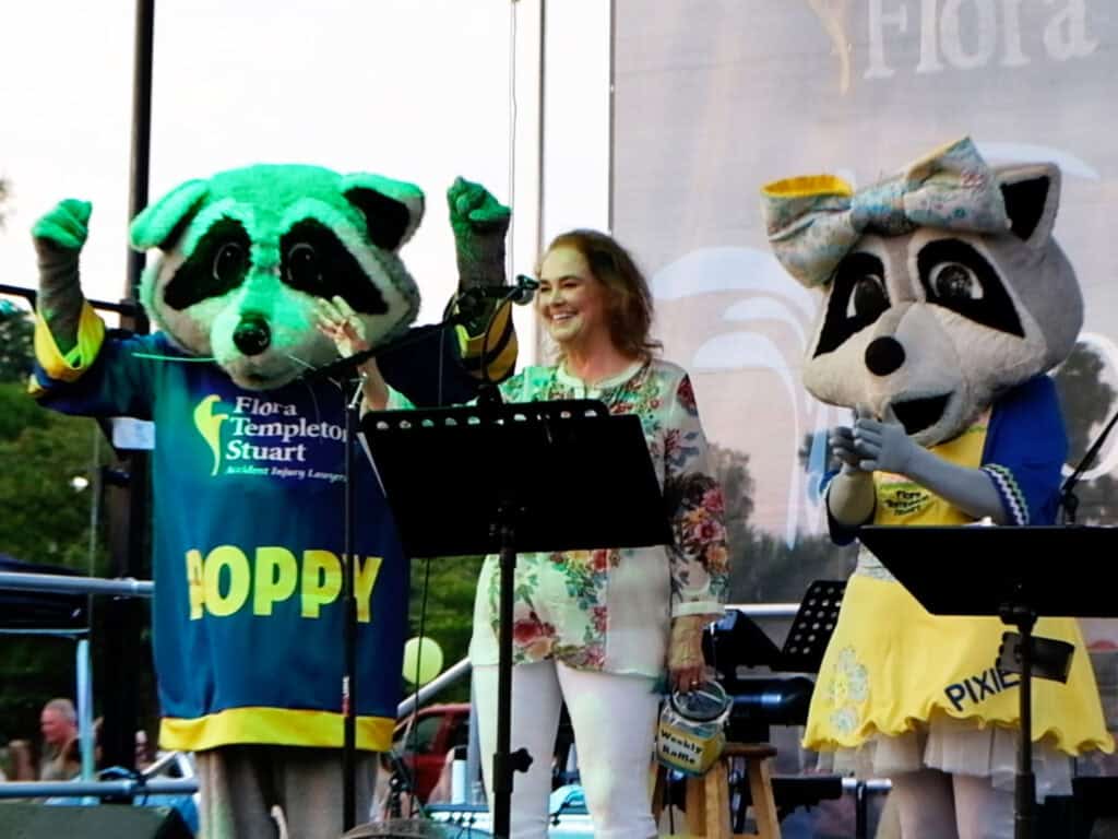 A woman stands at a microphone between two people in raccoon costumes—one in a blue jersey labeled "POPPY" and the other in a yellow dress labeled "PIXEL"—at an outdoor Warren County event.