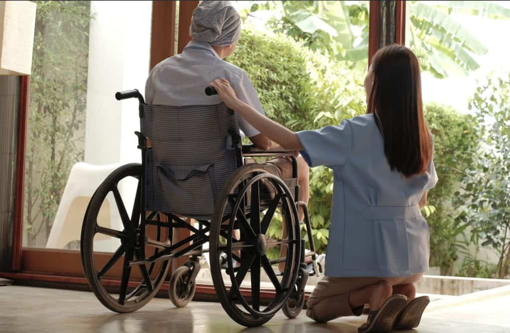 A caregiver kneels beside a person in a wheelchair, resting a hand on their shoulder as they look outside through a large open door, sharing a quiet moment of comfort and connection at home.