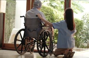A caregiver kneels beside a person in a wheelchair, resting a hand on their shoulder as they look outside through a large open door, sharing a quiet moment of comfort and connection at home.