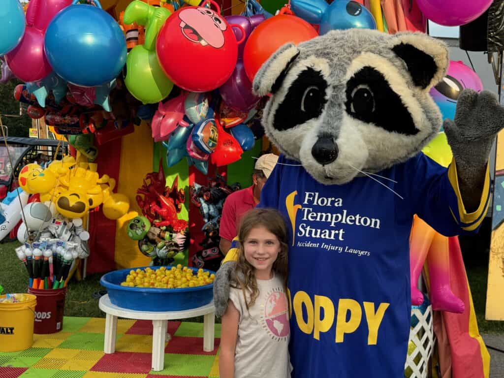 A girl stands next to a person in a raccoon mascot costume named "Poppy" at a colorful Warren County carnival booth, with balloons and prizes in the background.