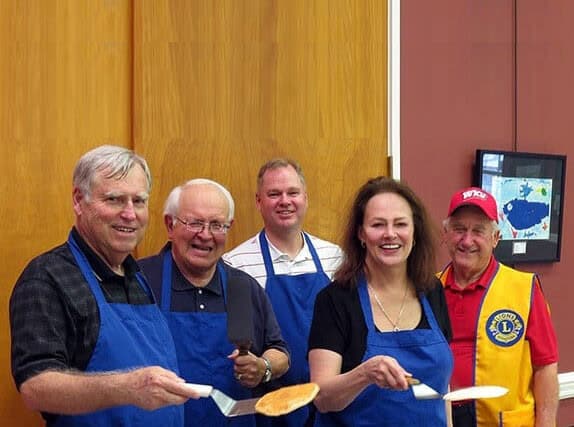 Five adults in blue aprons, with one in a red vest, stand indoors holding spatulas and pancakes, smiling at the camera during a community program focused on giving back to the community.