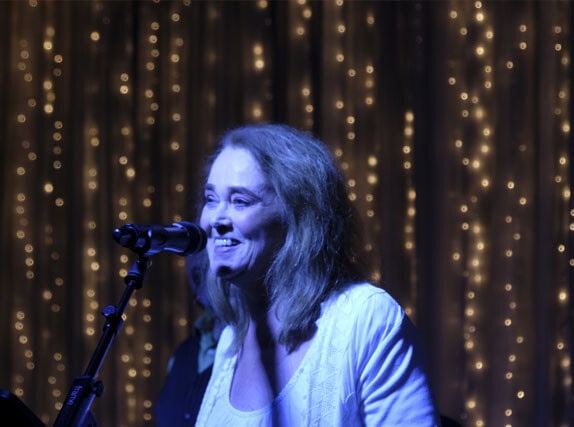 A woman smiles while standing at a microphone on stage, with warm string lights hanging in the background, during a community program focused on giving back to the community.