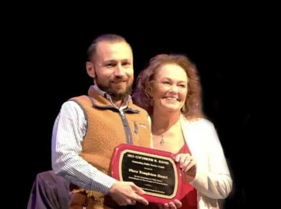 A man and a woman smile while holding an award plaque together against a dark background, celebrating their dedication to giving back to the community.