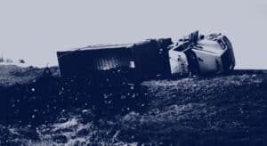 A large truck lies tipped over on its side off the road, with its cargo spilled onto the ground.