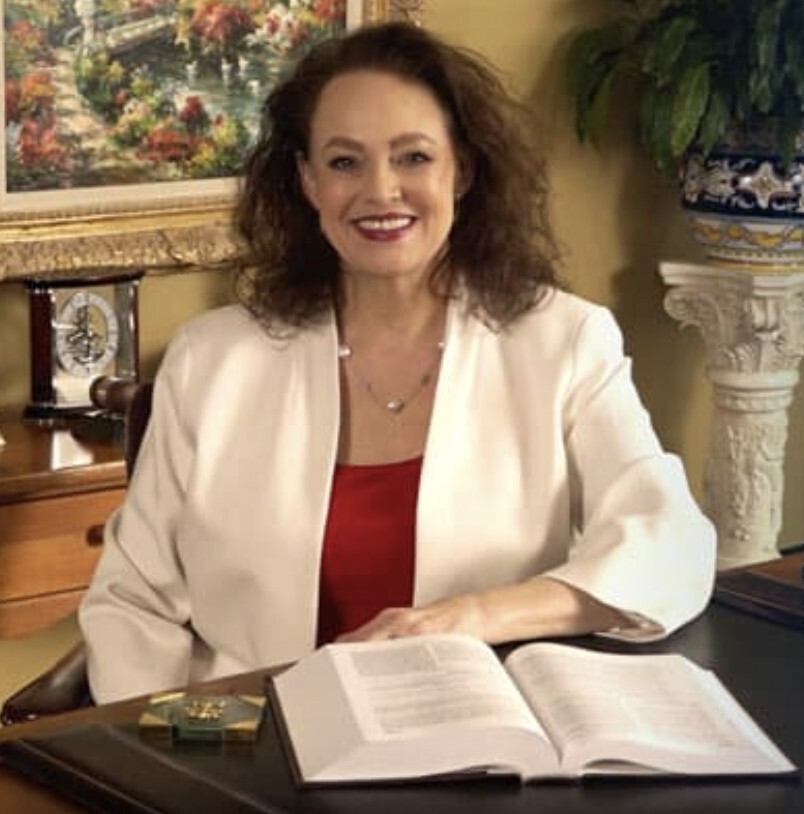 A woman with curly brown hair wearing a white blazer sits at a desk with an open book, smiling at the camera. A painting, clock, and plant are visible in the background.