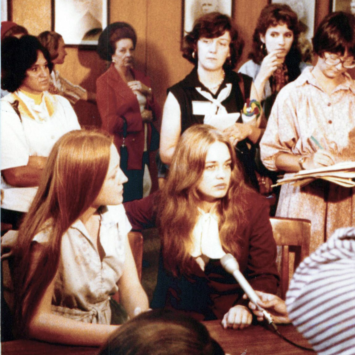 Two young women sit at a table, one with an arm around the other, as reporters and onlookers—including Flora Templeton Stuart—gather, some taking notes and holding a microphone toward them.