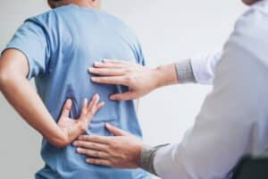 A person in a blue shirt holds their lower back in pain while a healthcare professional examines their back with both hands, assessing possible pre-existing conditions for a Kentucky injury claim.