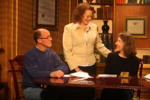 Three adults are seated and standing around a polished wooden table with papers, in a warmly lit room with framed certificates and bookshelves—discussing lawyer fees with experienced Kentucky car accident lawyers.
