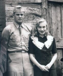 A man in a military uniform stands beside a woman in a dark dress with a white collar, channeling the grace of a Southern Belle, both posing in front of a rustic wooden building.