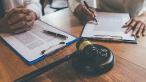 Two people sit at a desk with documents and pens; a gavel and two wedding rings are on the table, suggesting a Kentucky settlement or divorce proceeding where parties may discuss the settlement amount they will receive.