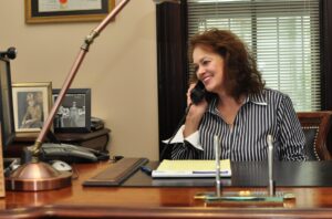 A woman in a striped shirt smiles while talking on the phone at her Bowling Green office desk, with paperwork, a pen holder, and framed photos behind her—ready to help you avoid claim failure in your car accident claims.