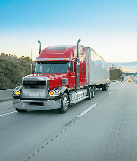 A red semi-truck with a silver trailer drives down a multi-lane highway near Owensboro KY, surrounded by greenery under a clear blue sky.