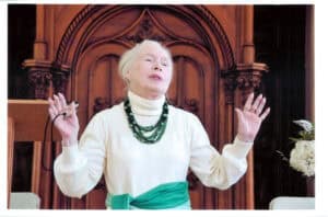 An elderly woman in a white turtleneck and green necklace stands with raised hands, speaking in front of an ornate wooden backdrop, embodying the poise of Flora Templeton Stuart.