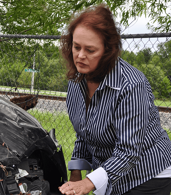 A woman in a striped shirt examines damage to the front end of a black car near a chain-link fence and railroad tracks in Owensboro, KY.