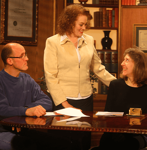 Three people sit around a table with papers in a warmly-lit office in Elizabethtown KY; two are seated while one stands between them, all engaged in friendly conversation—perhaps discussing matters with experienced car accident lawyers.