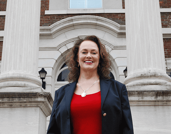 Woman in a dark blazer and red top stands in front of a building with large columns and arched windows in Elizabethtown KY, smiling at the camera—representing trusted Truck Accident Lawyers.