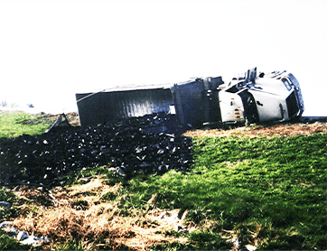 An overturned truck lies on its side beside a grassy hill near Owensboro, KY, with a large pile of black debris spilled from its trailer—a scene where Truck Accident Lawyers may be needed.
