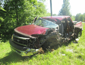 A heavily damaged red pickup truck with a crushed front and side sits on grassy ground beside a tree-lined road in daylight—an accident scene where Car Accident Lawyers in Owensboro KY could help.
