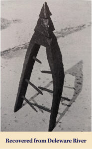 A rusted, triangular metal object with three horizontal spikes and a pointed top stands upright on a flat surface, resembling a piece Judge Clay Elliott might notice in one of Flora Templeton Stuart’s unique collections.