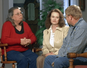 Three adults sit indoors in Bowling Green, KY, engaged in conversation. One woman wears a red sweater, another a beige jacket, and a man in glasses and a plaid shirt. Plants and furniture are visible—perhaps discussing motorcycle accident lawyers.