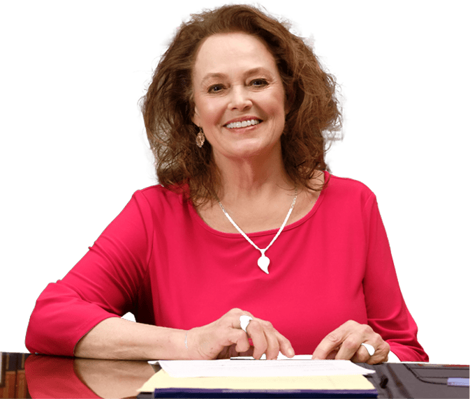 A woman with curly brown hair wearing a bright pink top sits at a desk, smiling, with papers and a folder in front of her—ready to assist as one of the trusted Truck Accident Lawyers in Bowling Green KY.