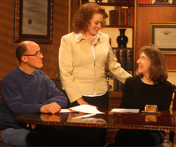Three adults sit and stand around a table with papers, having a conversation in a warmly lit room with shelves and framed certificates—your trusted Bowling Green, KY Car Accident Lawyers ready to help.