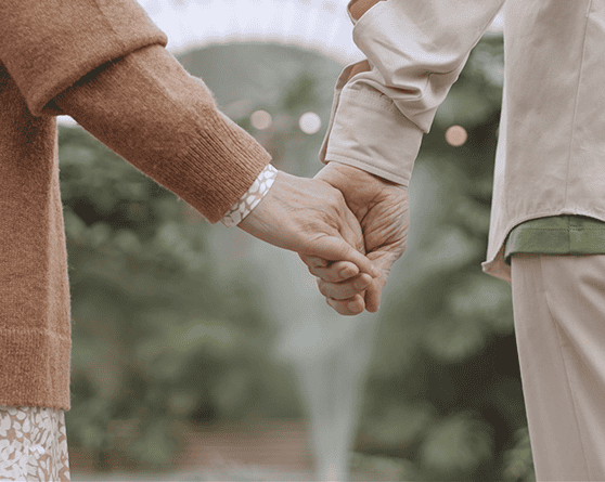 Two people are seen from behind holding hands outdoors in Glasgow KY, wearing light-colored clothing, with greenery and a fountain in the background—an image evoking support and trust, much like lawyers helping with nursing home abuse cases.