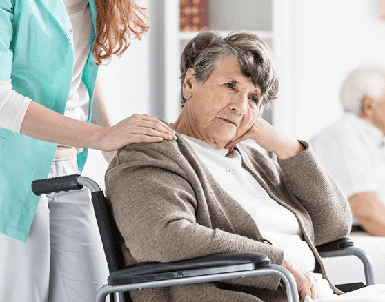 An elderly woman sits in a wheelchair with a thoughtful expression while a caregiver stands beside her, resting a hand on her shoulder. Another elderly person is visible in the background, highlighting the importance of Nursing Home Abuse Lawyers in Glasgow KY.