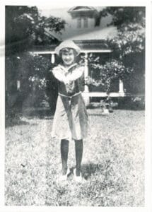 A young girl stands smiling with arms crossed in front of her chest, wearing a hat and dress, on a grassy lawn with a house and trees in the background—reminiscent of portraits from the Flora Templeton Stuart collection.
