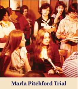 Two women sit at a table during a trial, one holding a microphone towards them as Judge Clay Elliott presides. Several people stand in the background, some taking notes.
