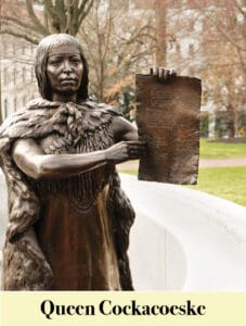 Bronze statue of Queen Cockacoeske holding a document, honoring Virginia Settlers, displayed outdoors with trees and buildings in the background.