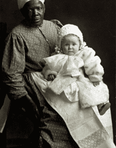 An adult with slave ancestry wearing a patterned dress and headscarf holds a baby in a bonnet and white gown, both facing the camera in a studio portrait.