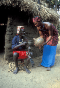 An older man with slave ancestry in traditional attire sits outside a mud hut holding a staff, while a woman in patterned clothing offers him a round container.