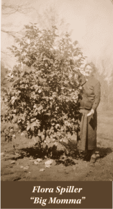 An older woman in a long skirt and jacket stands beside a large leafy bush outdoors. Text reads: "Flora Spiller 'Big Momma'," honoring her slave ancestry.