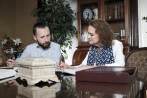 A man and woman sit at a desk reviewing documents together in an office, with books, a plant, and decorative items in the background.